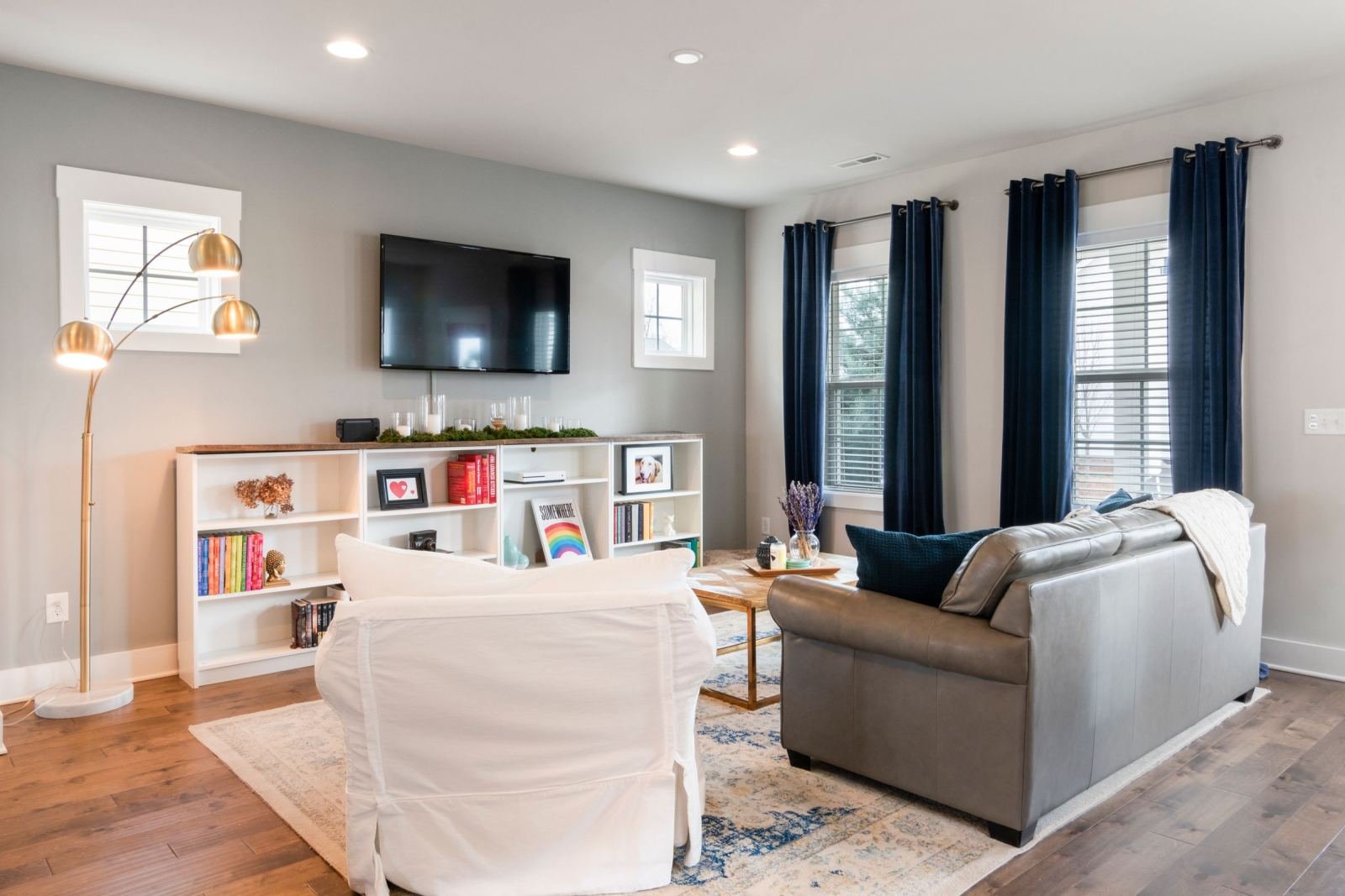 A bright living room with a grey sofa, white armchair, and a bookshelf under a wall-mounted TV