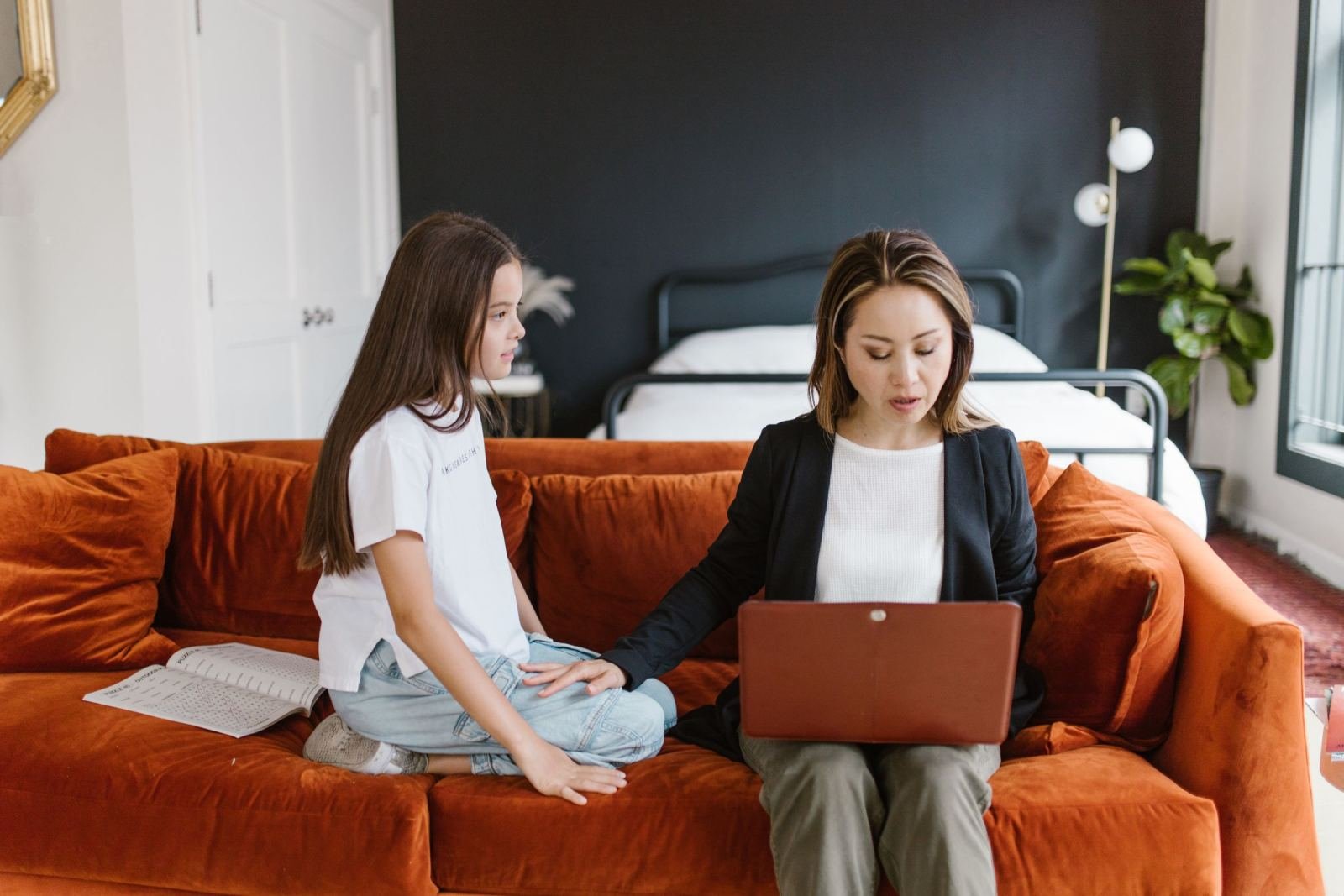 A mother and her daughter sit on an orange couch, checking a laptop.