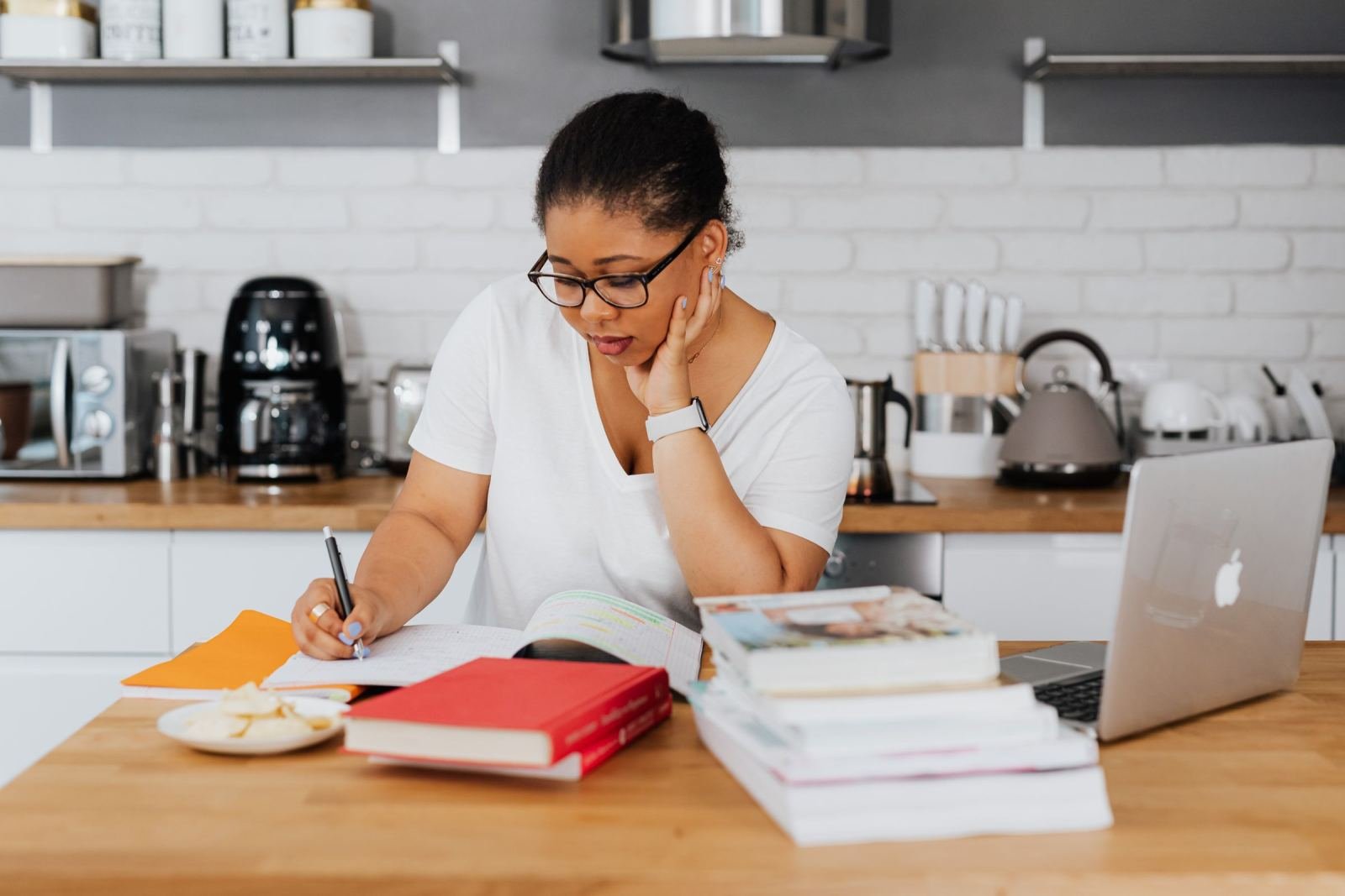 A woman wearing glasses writes in a notebook at a kitchen counter with a laptop