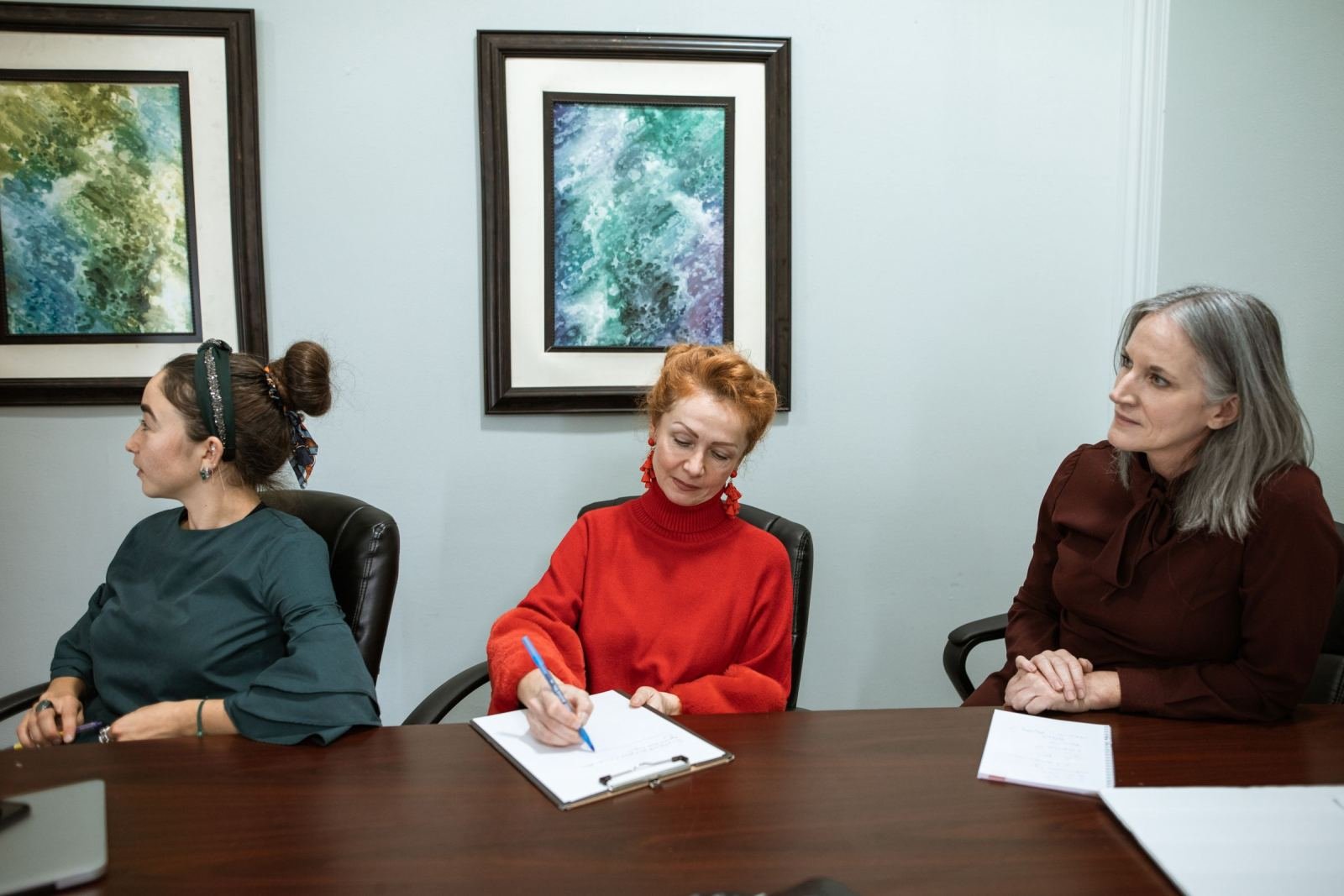 Three women sit around a wooden table in a bright office with artwork on the walls