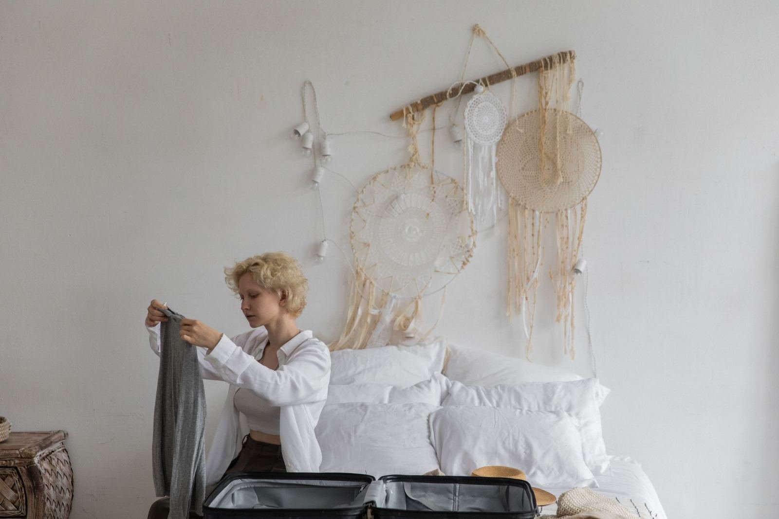 A woman holds a grey piece of clothing over an open suitcase on a bed