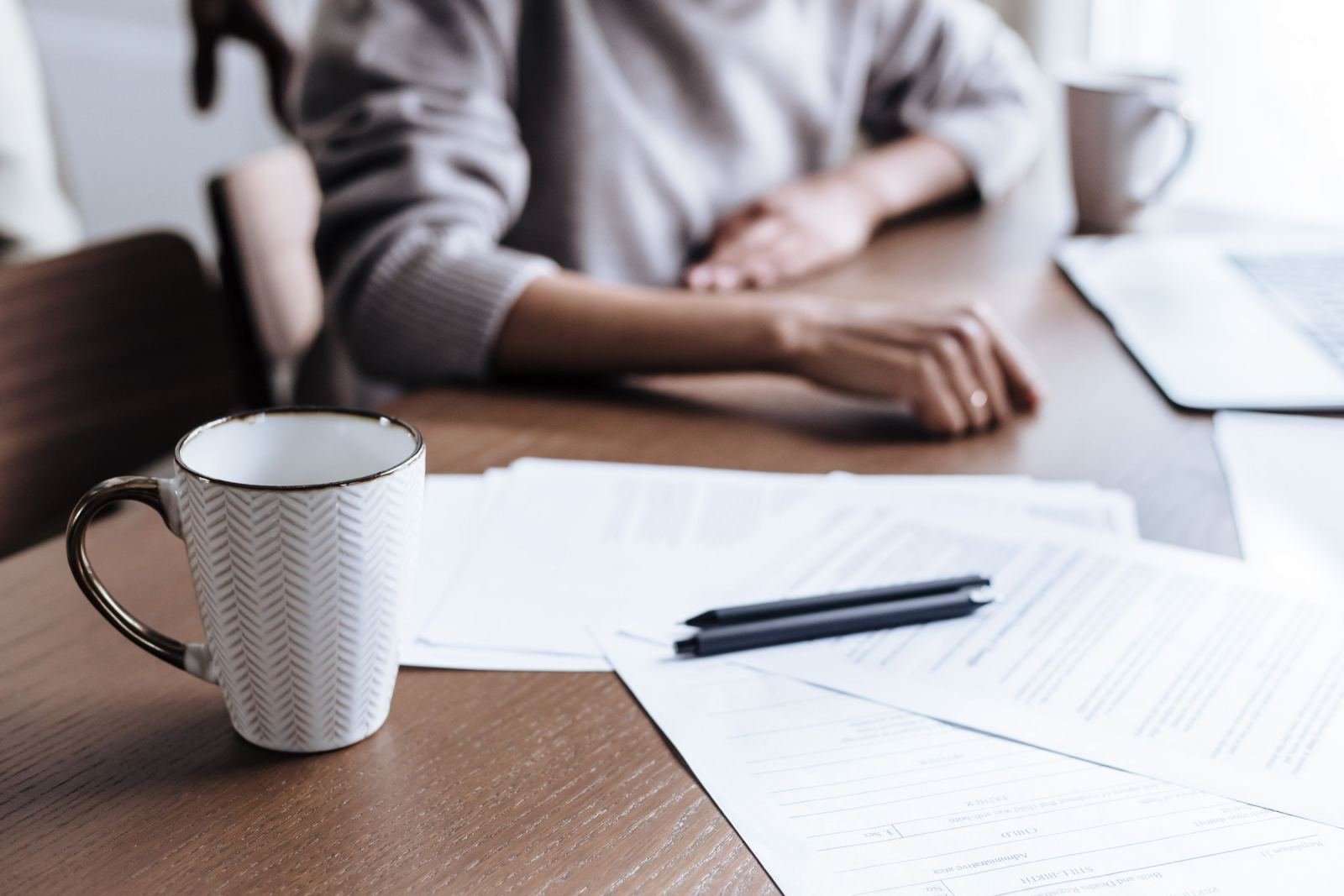 A person sits at a wooden table with printed documents, a pen, and a mug