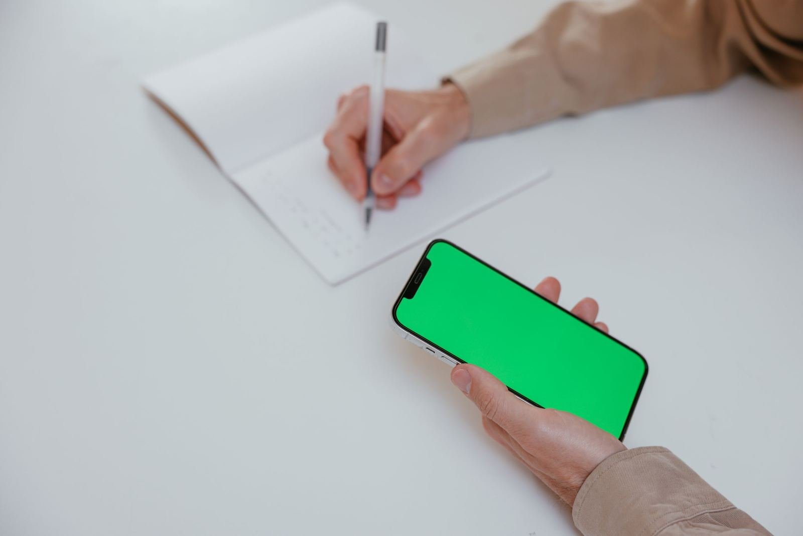 A person uses a notebook and a phone with a green screen.