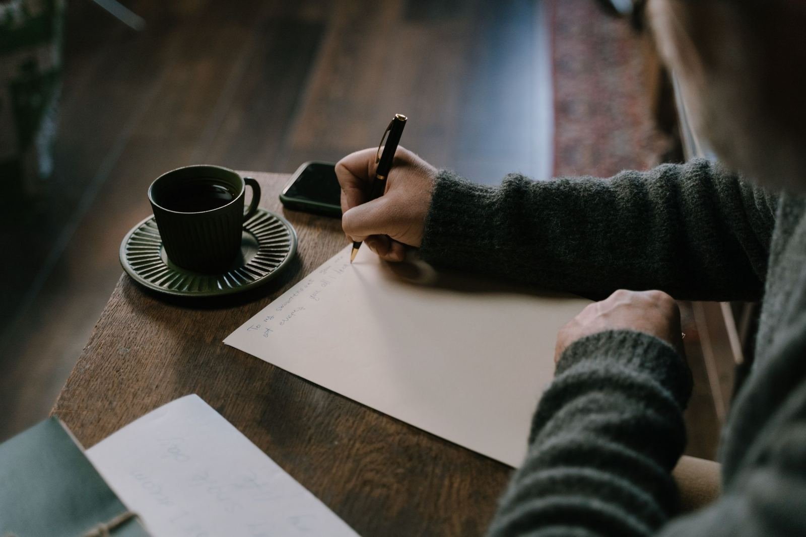 A person in a green sweater writes on a piece of paper at a wooden table