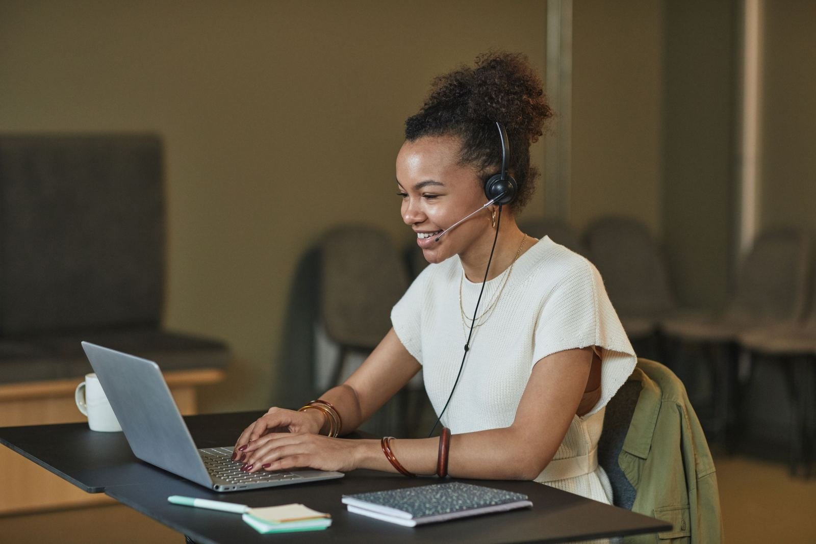 A smiling woman uses a laptop at her desk while wearing a headset.