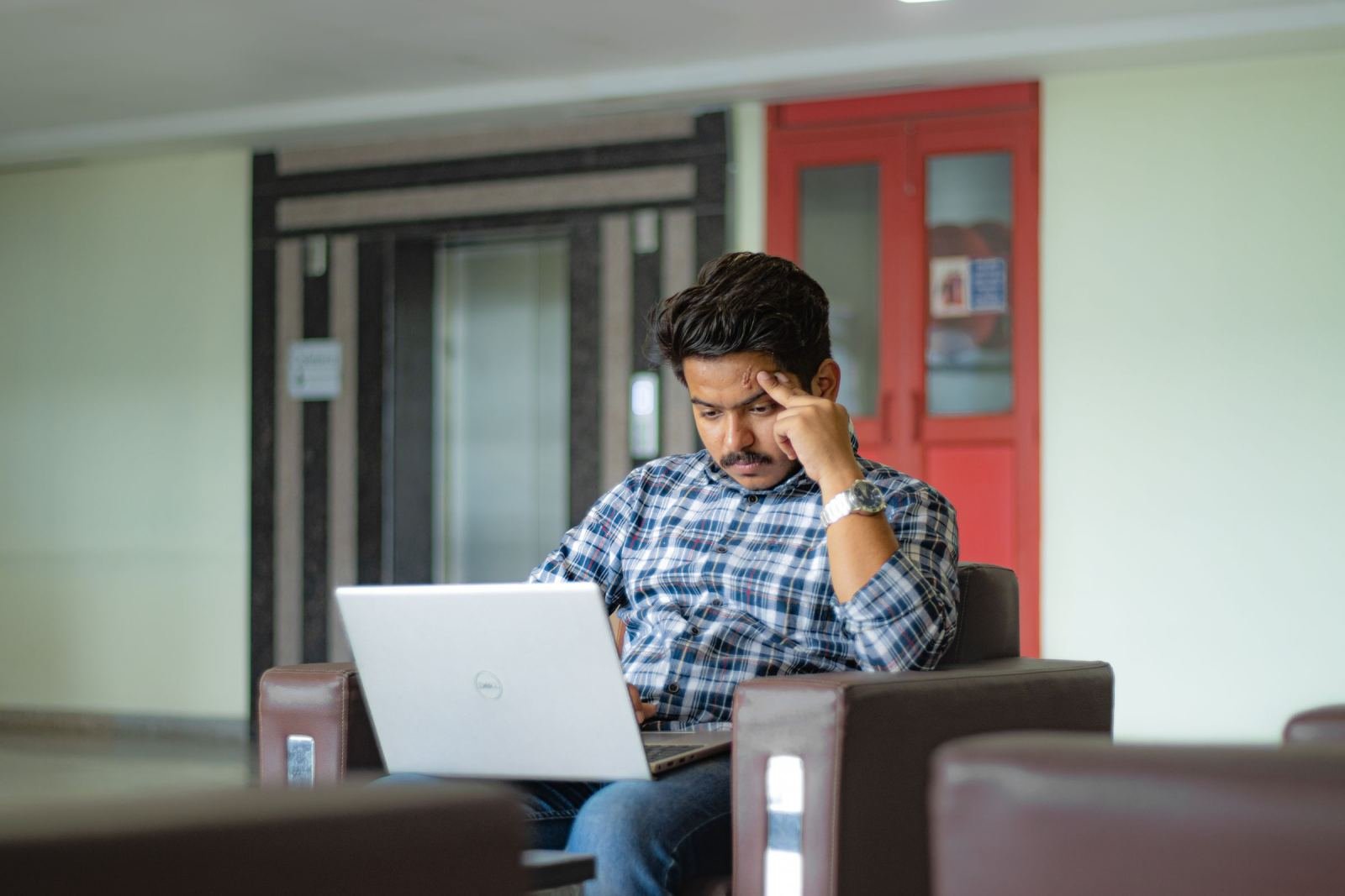 A man in a plaid shirt sits in an armchair using a laptop with his hand on his forehead