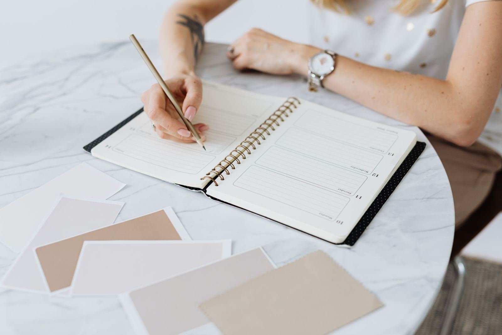 A person writes in a spiral planner on a marble table with color swatches
