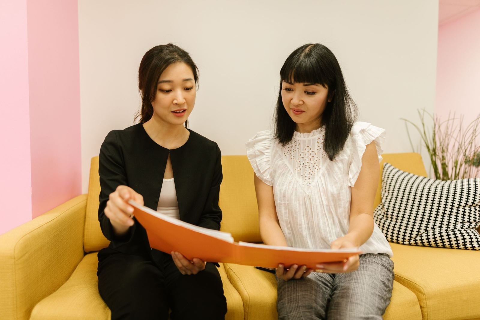 Two women sit on a yellow sofa reviewing documents in an orange folder