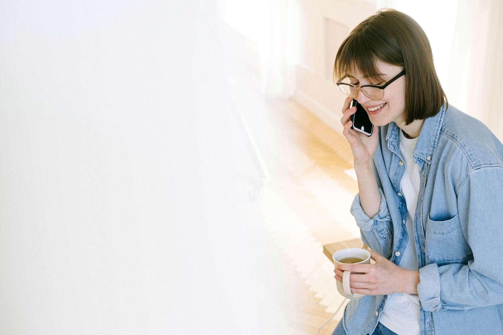A smiling woman with glasses talks on a mobile phone while holding a mug