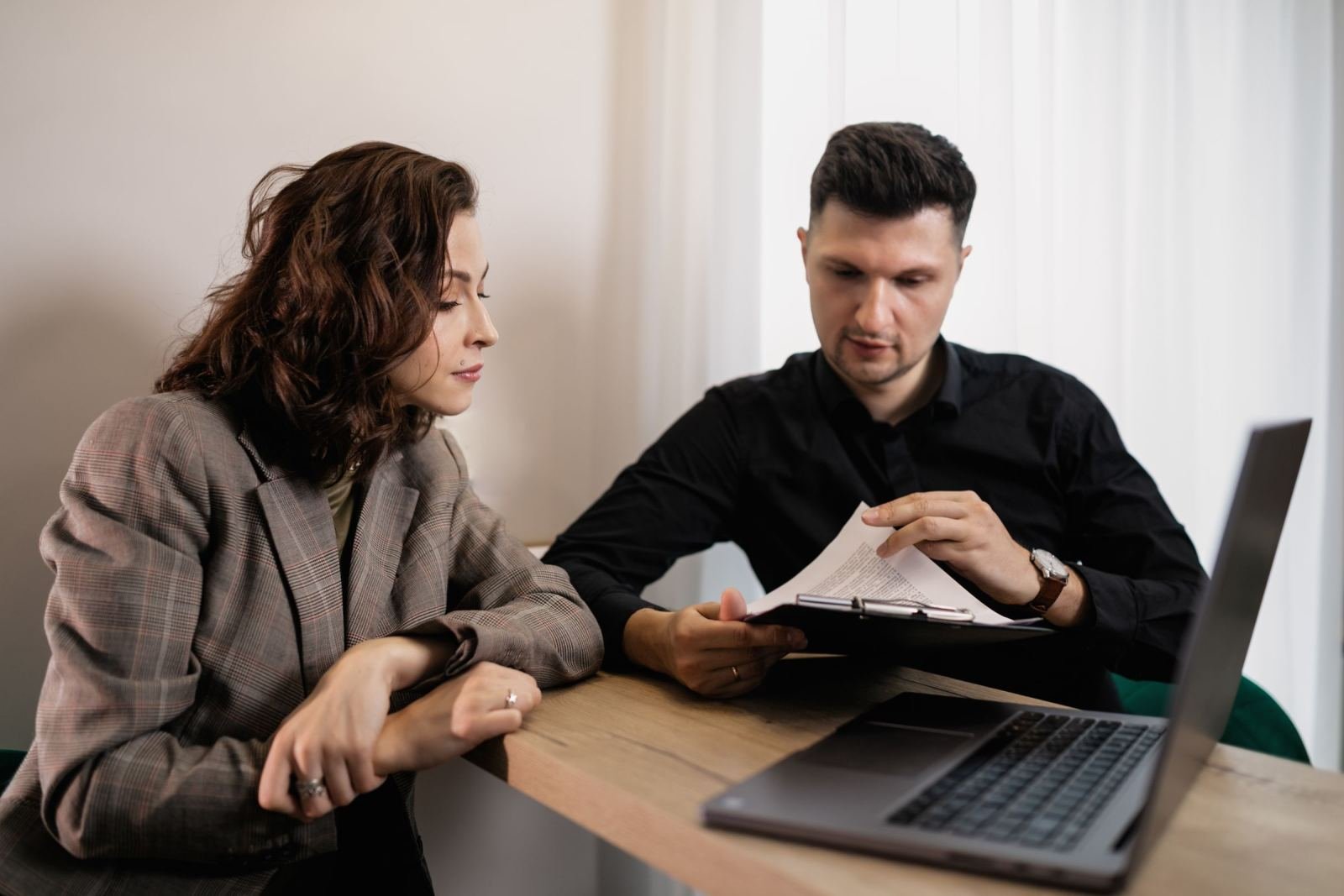 A man and woman sit at a table looking at a laptop and a clipboard
