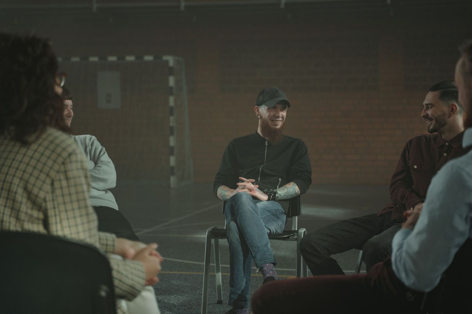 A group of people sit in a circle of chairs in a dim room for a group discussion