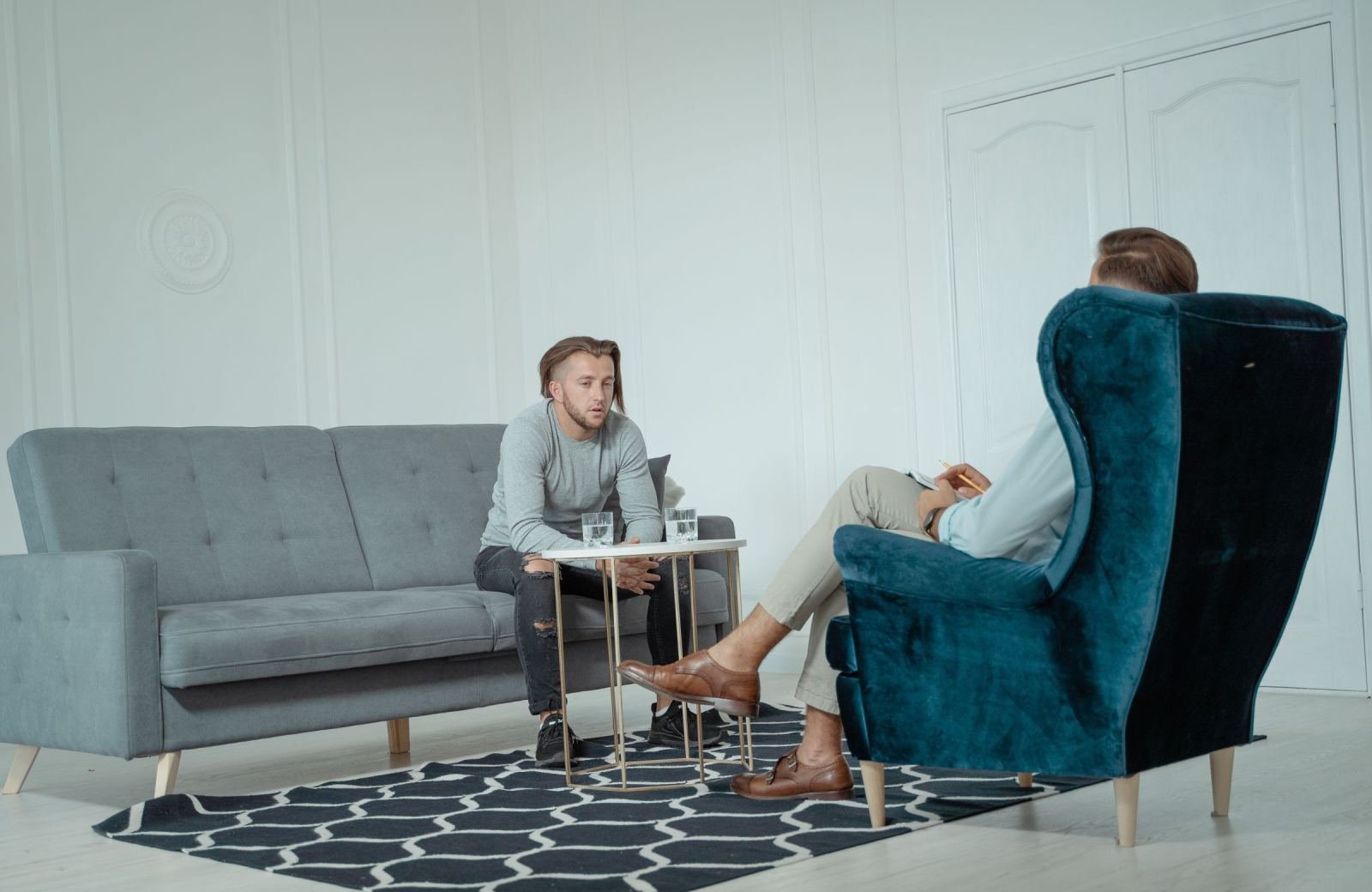 A man sits on a sofa talking to a professional in a blue velvet armchair