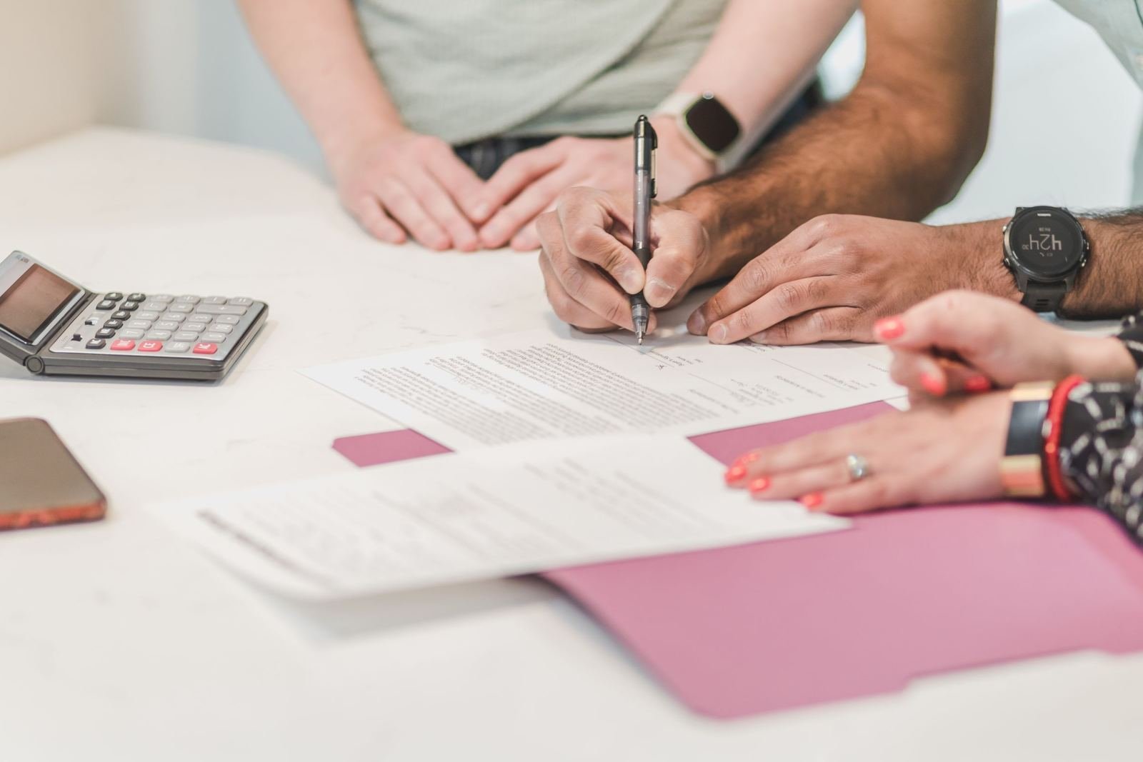 A person signs a document on a white table next to a calculator and a purple folder