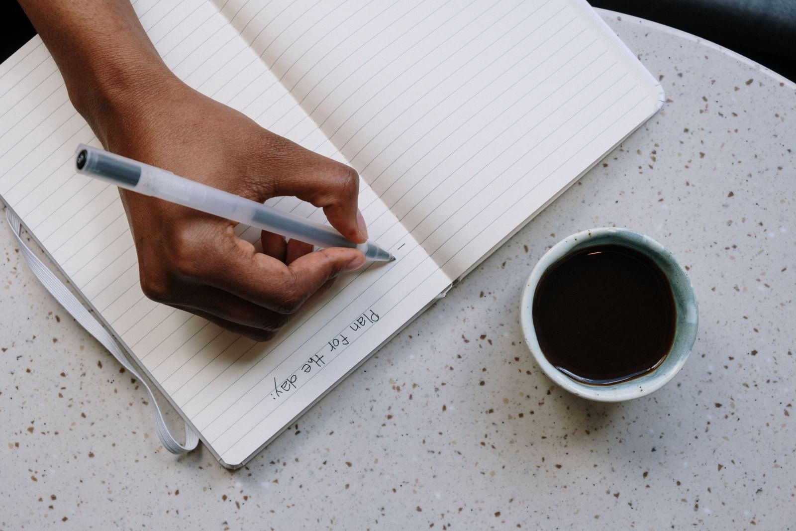 A person writes a daily plan in a notebook next to a cup of coffee