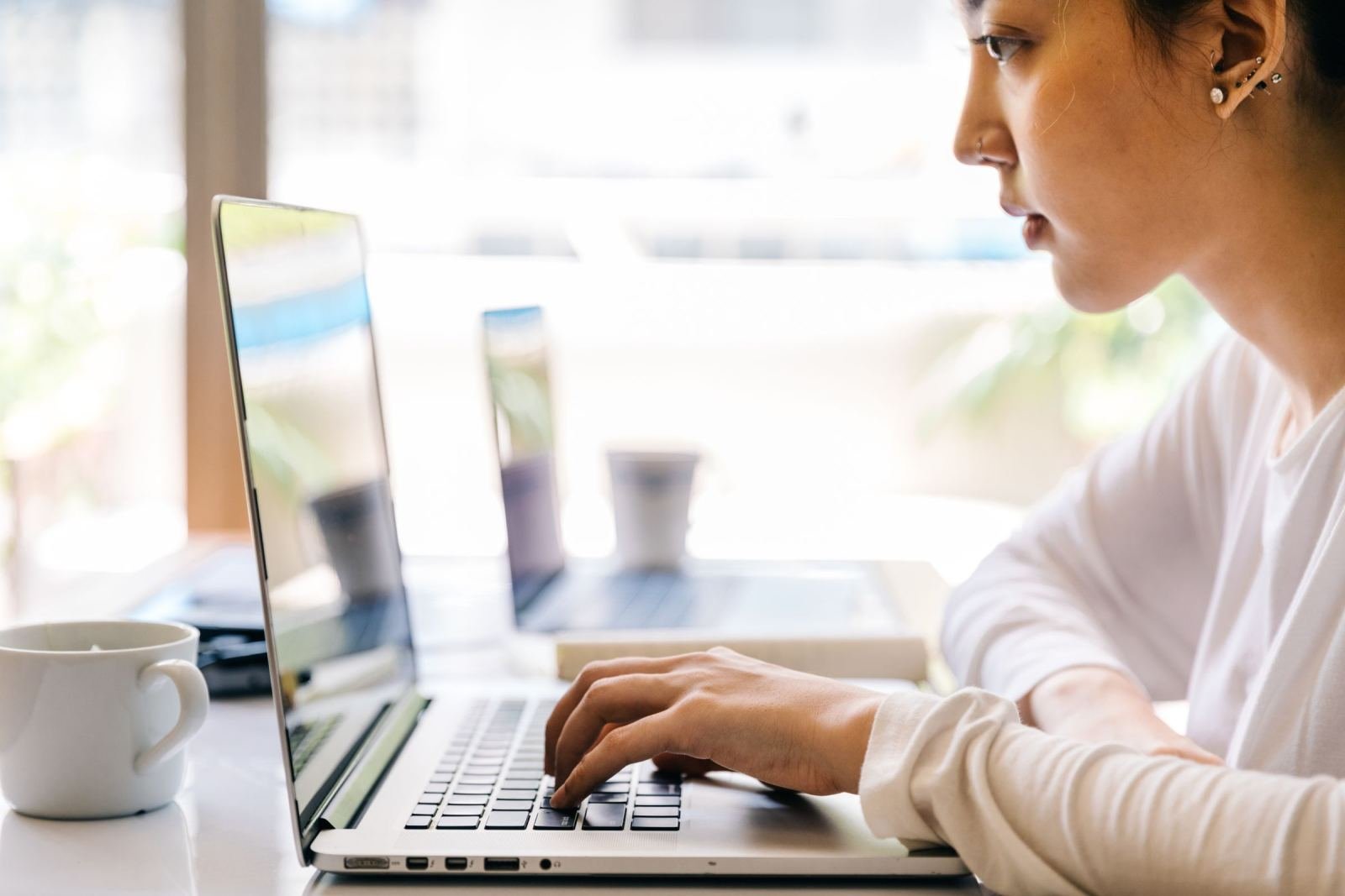 A woman works on a laptop at a bright desk with a white mug close by