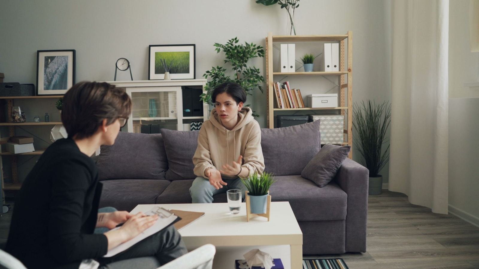 A teen sits on a grey sofa while a clinician in a black blazer talks with them.