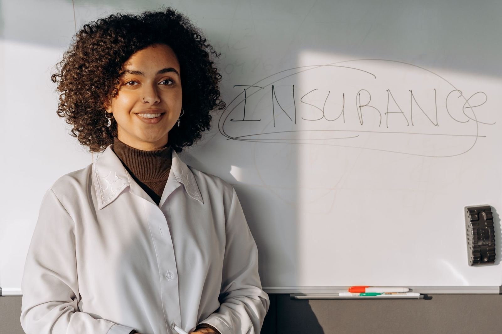 A woman in a white lab coat stands in front of a whiteboard with the word Insurance written on it