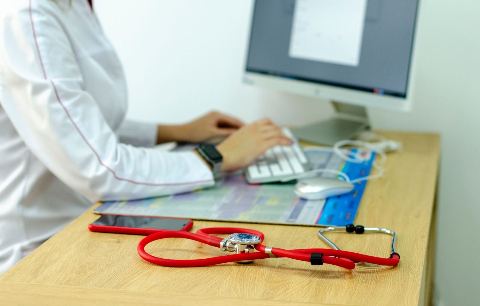 Image shows a healthcare provider in a white coat typing at a desk with a red stethoscope in the foreground.