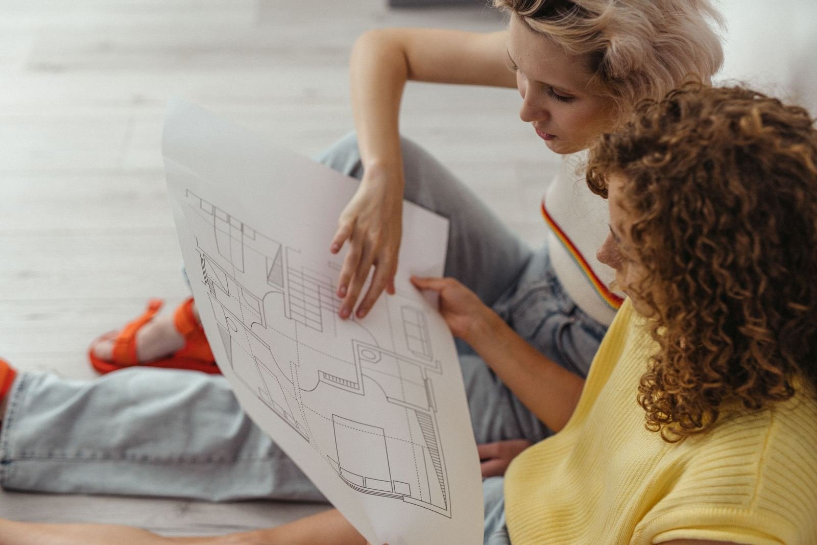 Two women sit on the floor reviewing a large architectural blueprint