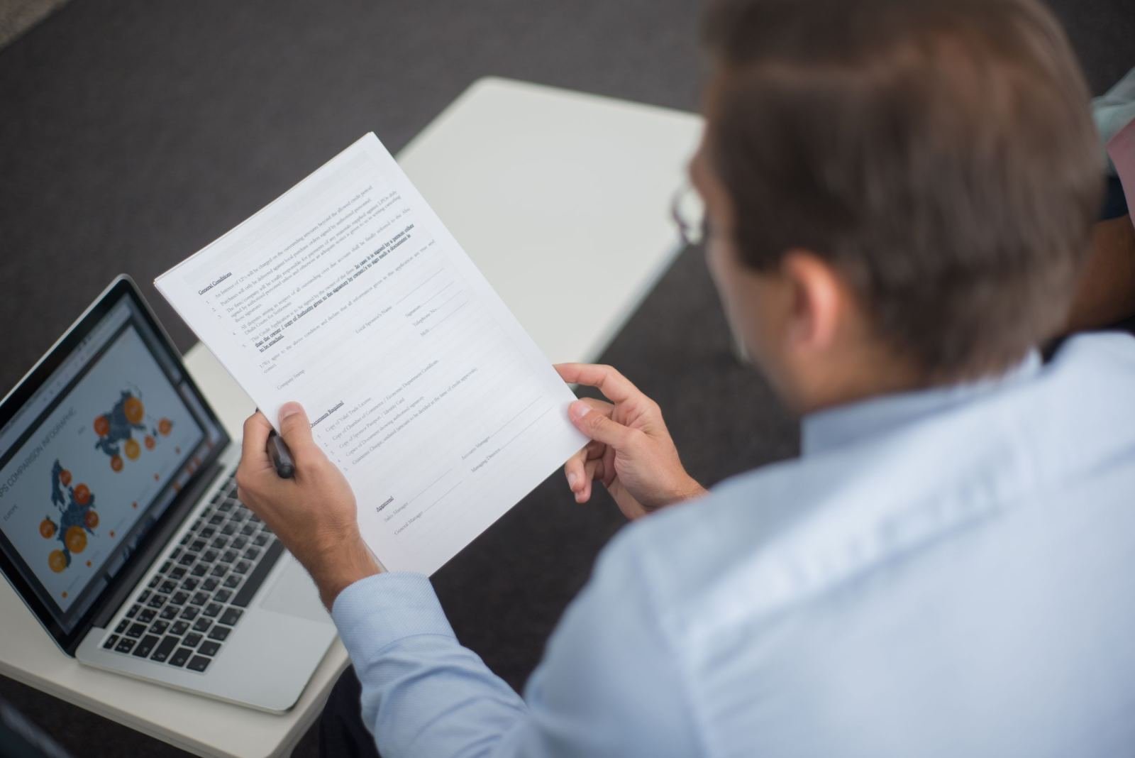 A man in a blue shirt reviews a printed document next to a laptop