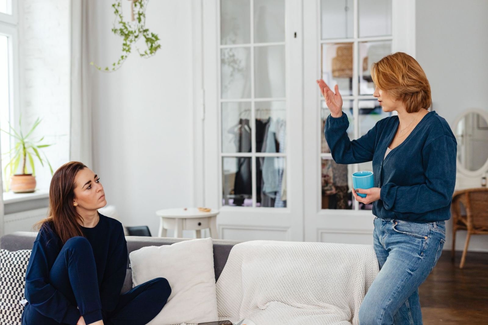 A woman sitting on a sofa looks up at another woman who is standing and gesturing