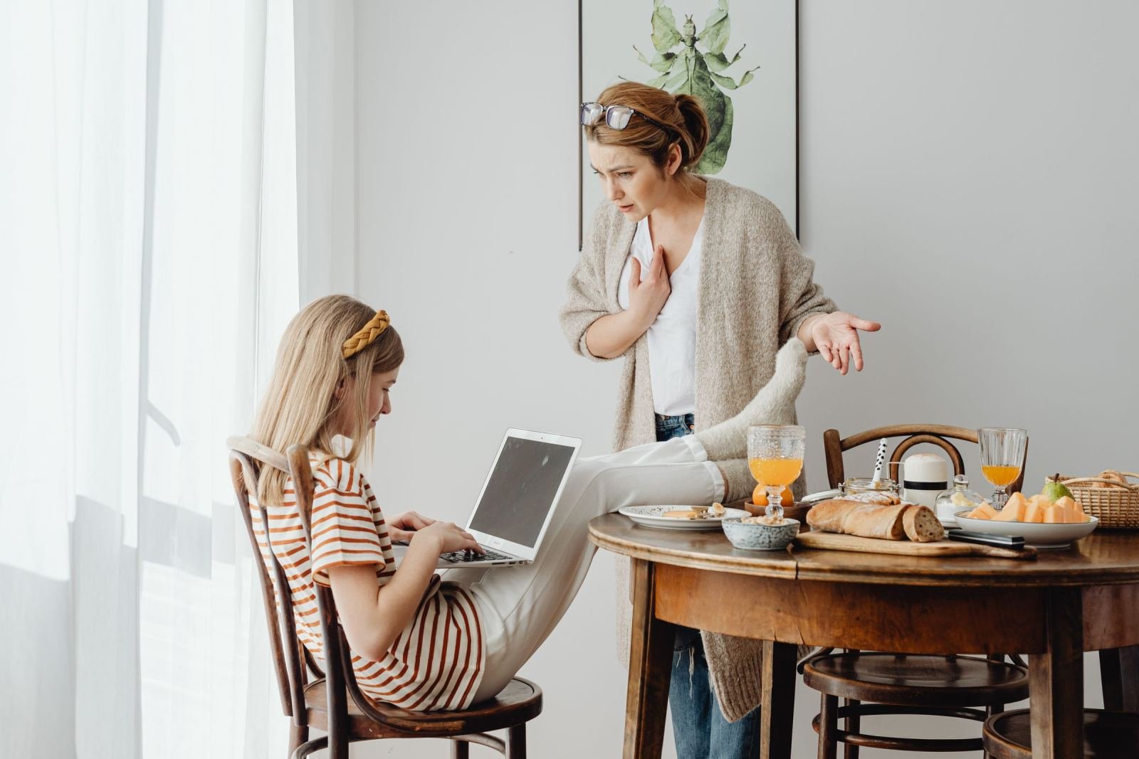A woman uses hand signals while speaking to a younger woman at a dining table with a laptop.
