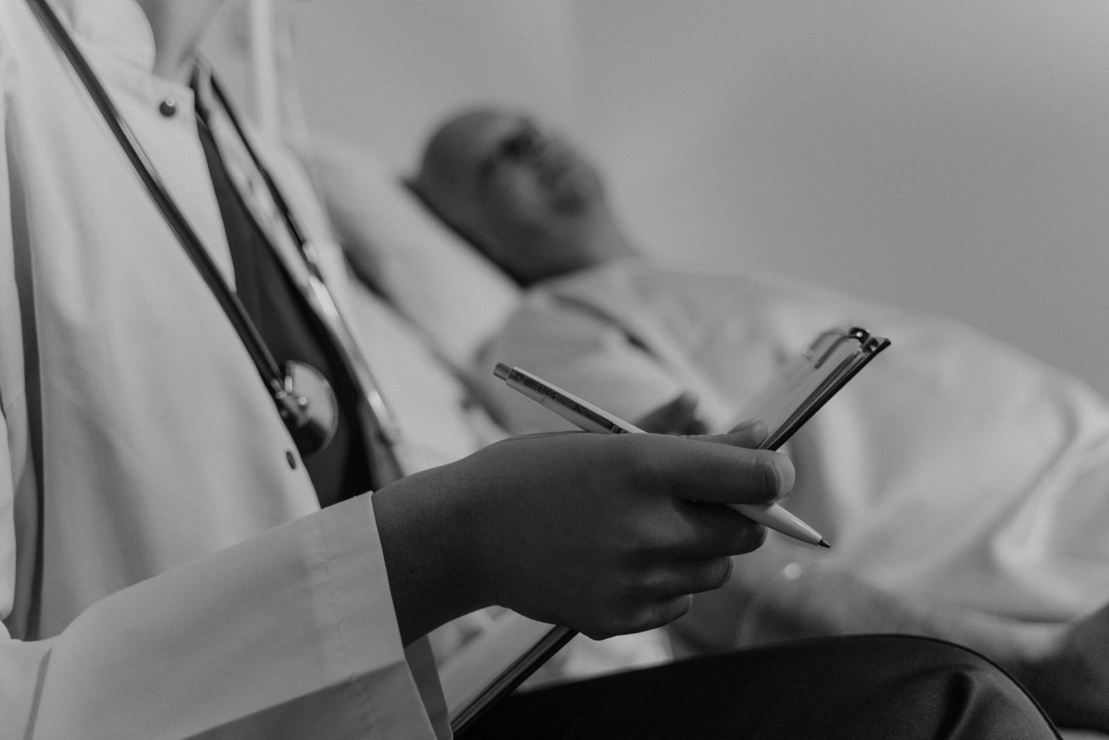 A clinician in a white coat holds a clipboard while a patient lies in the background