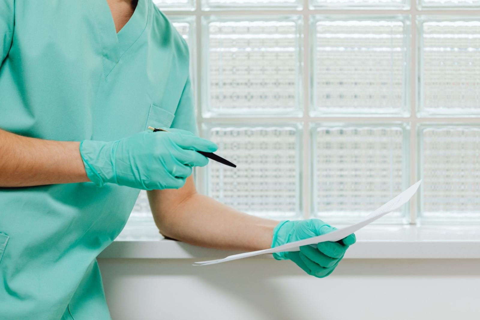 A medical professional wearing green scrubs and gloves holds a document and a pen.