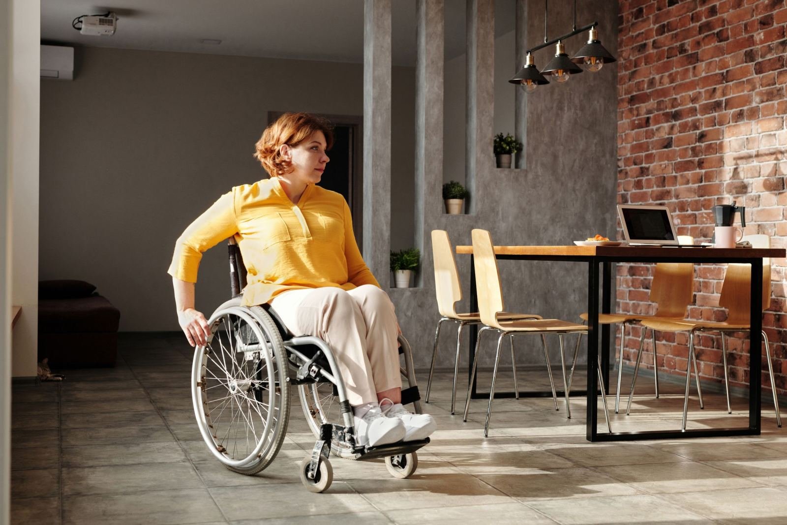 A woman in a yellow shirt sits in a wheelchair in a room with a brick wall