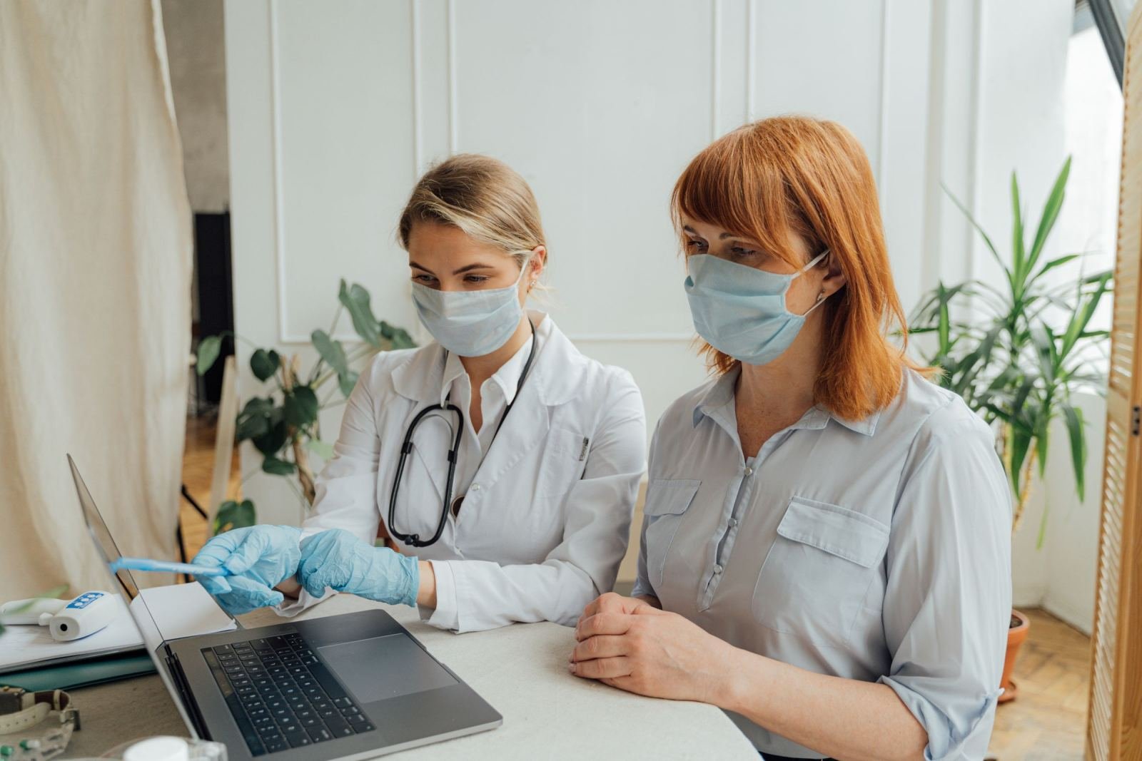 A clinician in a white coat and mask points to a laptop screen while talking to a patient