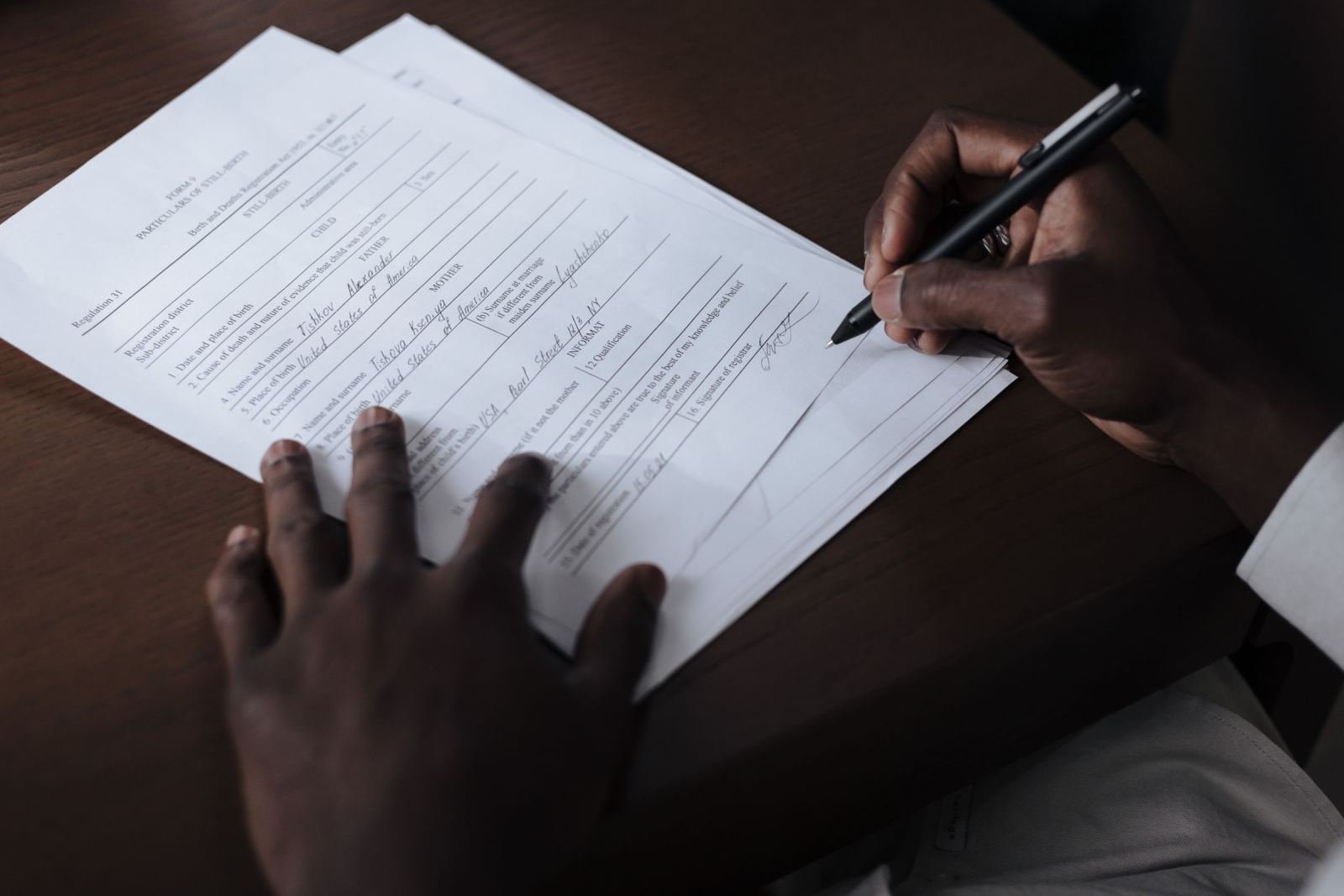 A person filling out a printed form with a black pen on a dark desk