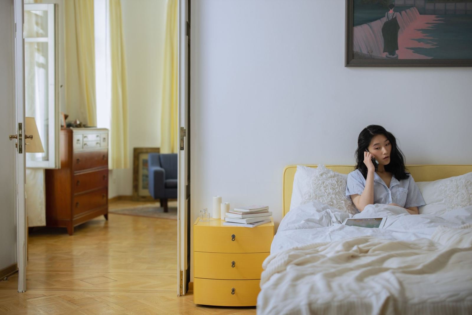 A woman sits in bed talking on a phone in a sunlit room