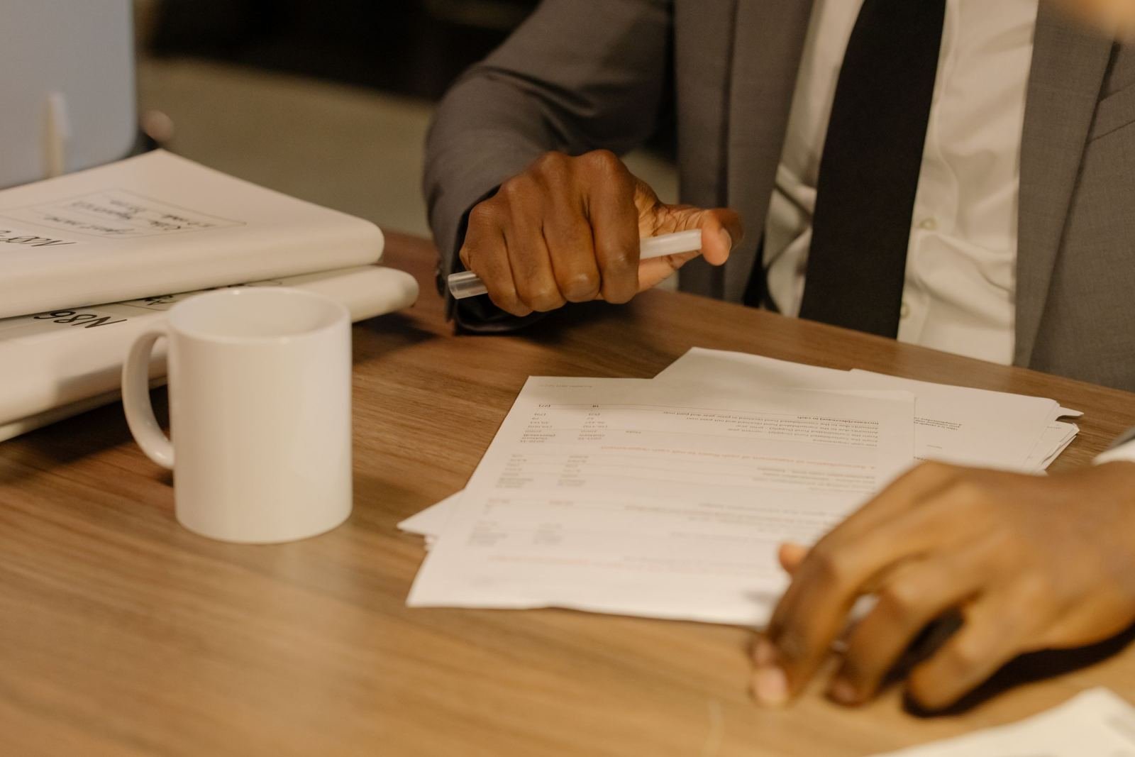 A person in a suit holds a pen over paperwork on a wooden desk