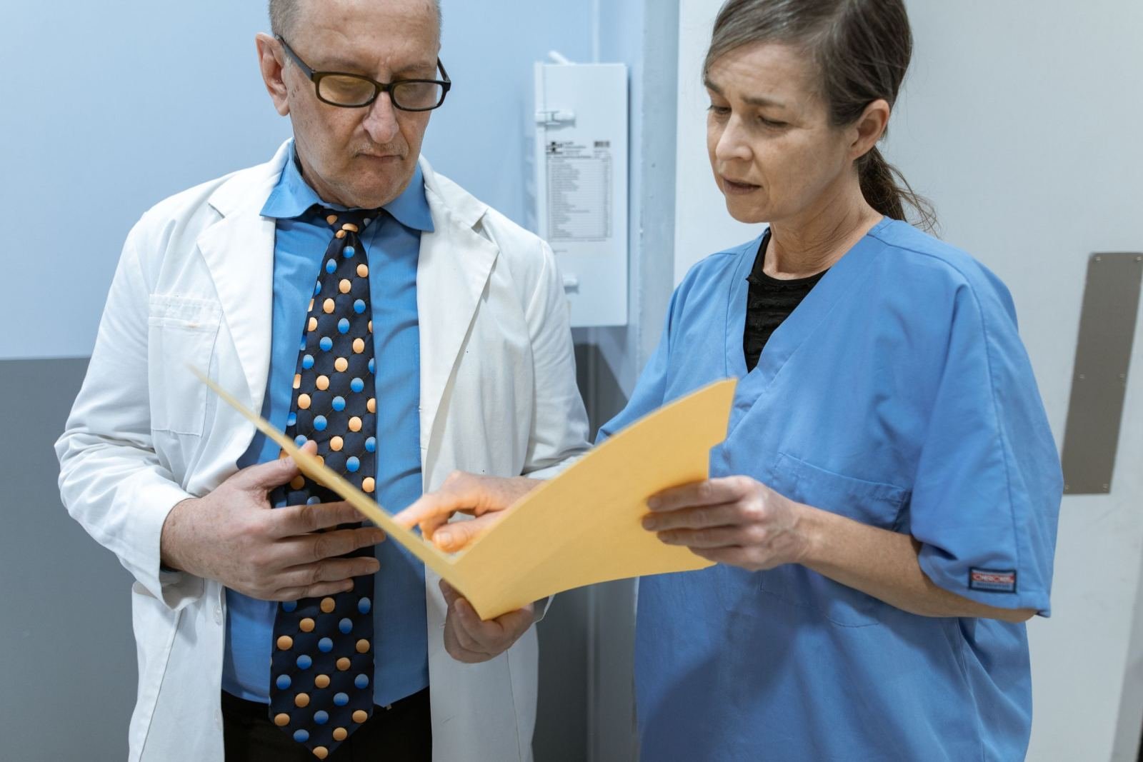 A doctor in a white coat and a nurse in blue scrubs review a yellow folder