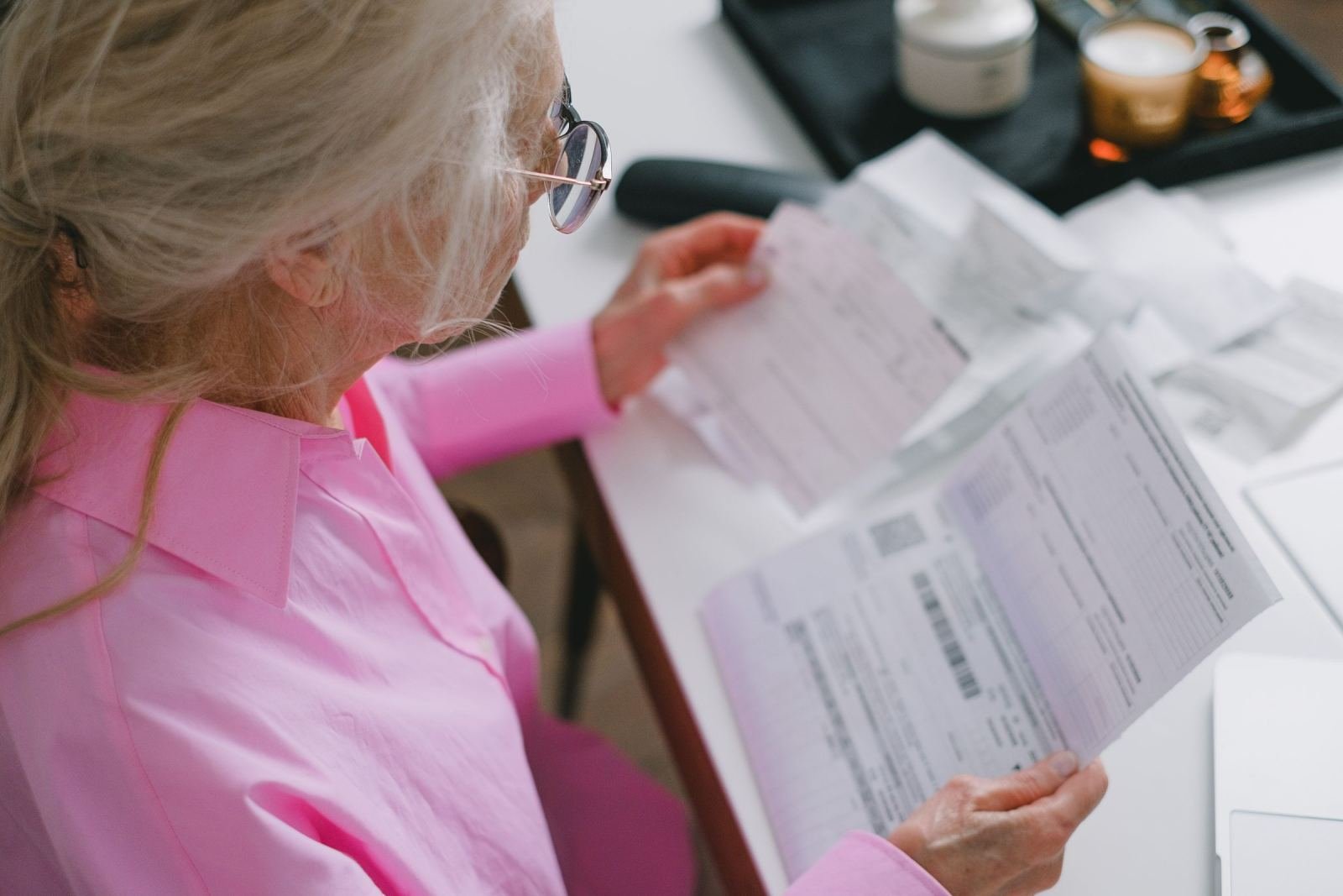 A white-haired woman in a pink top looks over paper files on a light desk.