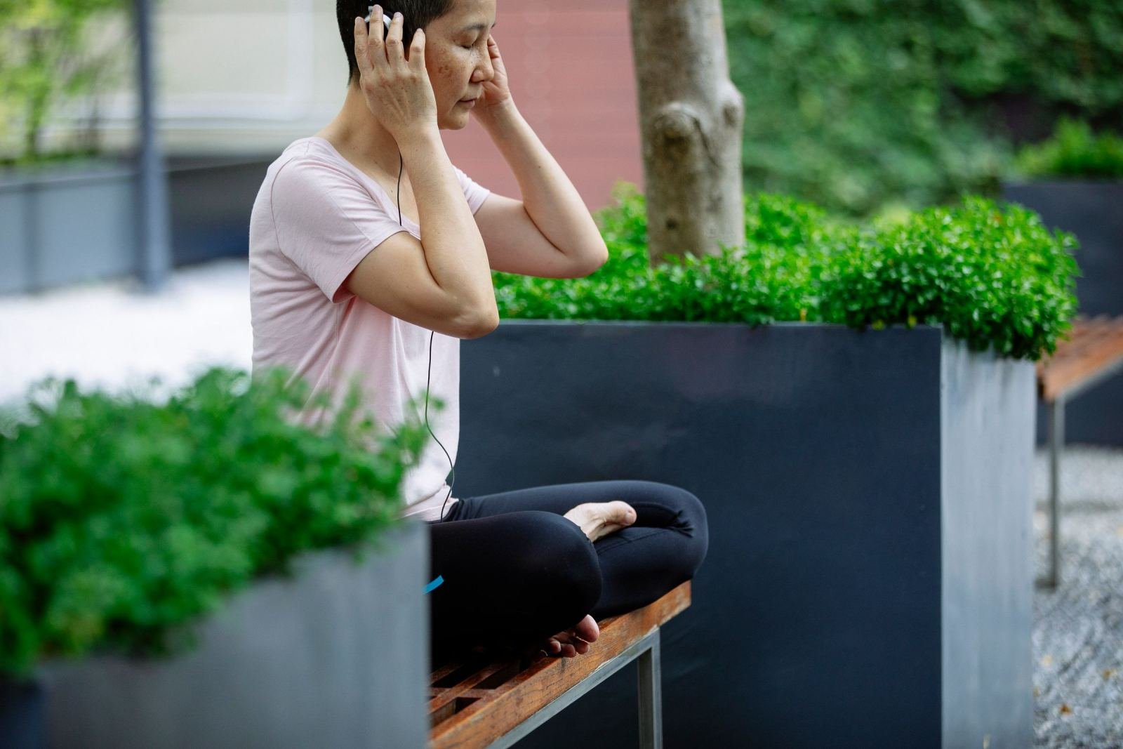 A woman sits cross-legged on a park bench with her eyes closed and earbuds in