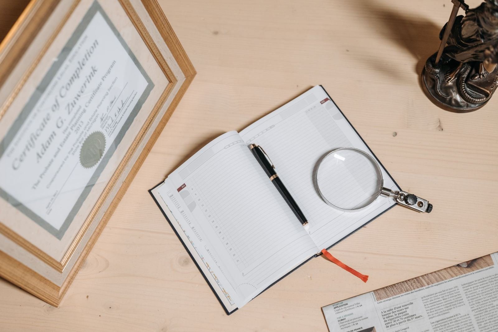 A framed certificate, an open notebook with a pen, and a magnifying glass on a wooden desk