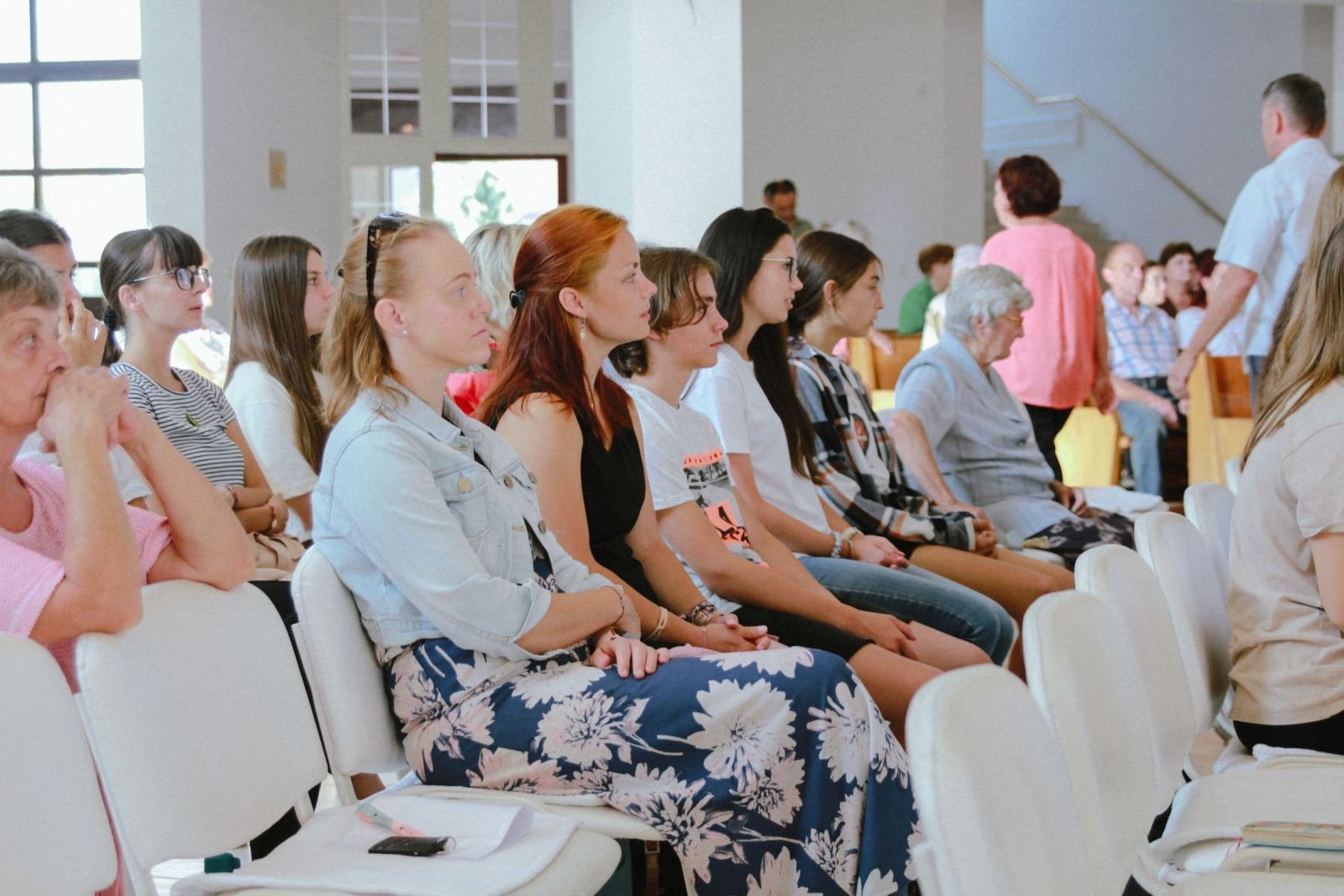 A group of people sit in white chairs and listen during a meeting in a bright room
