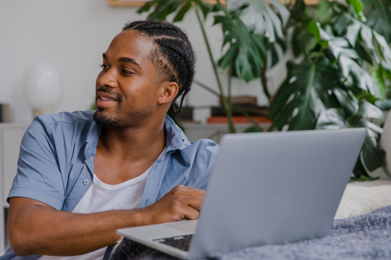 A man with braided hair sits at a table with a laptop and looks to the side