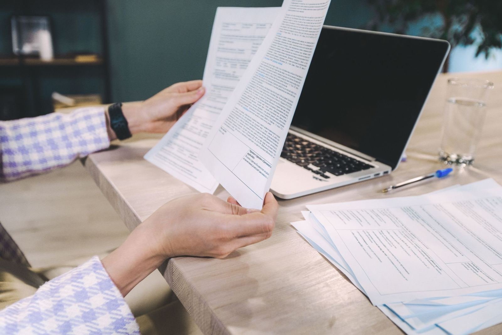 A person reviews printed documents at a table next to an open laptop