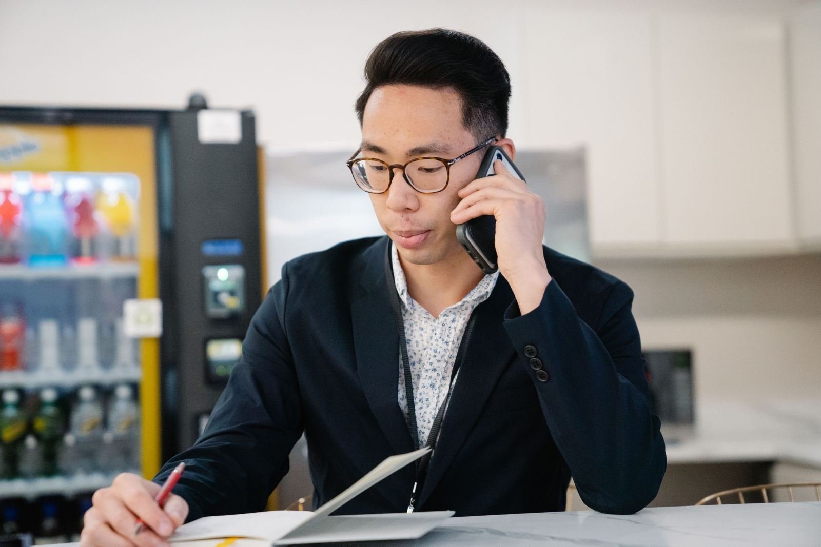 A man in a blazer talks on a phone while writing in a notebook at a desk