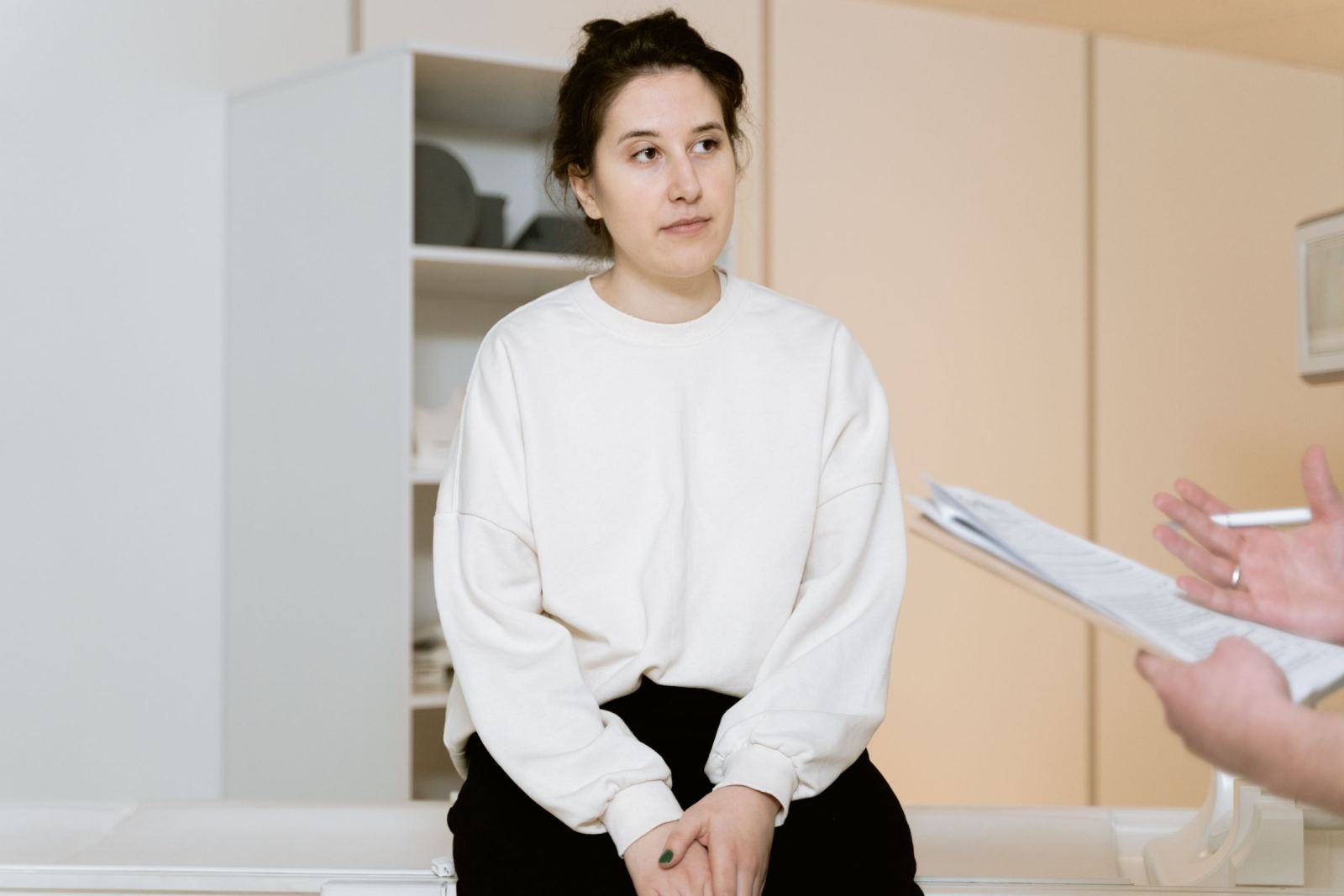 A clinician stands beside an exam table, holding a clipboard, while a woman sits on it.