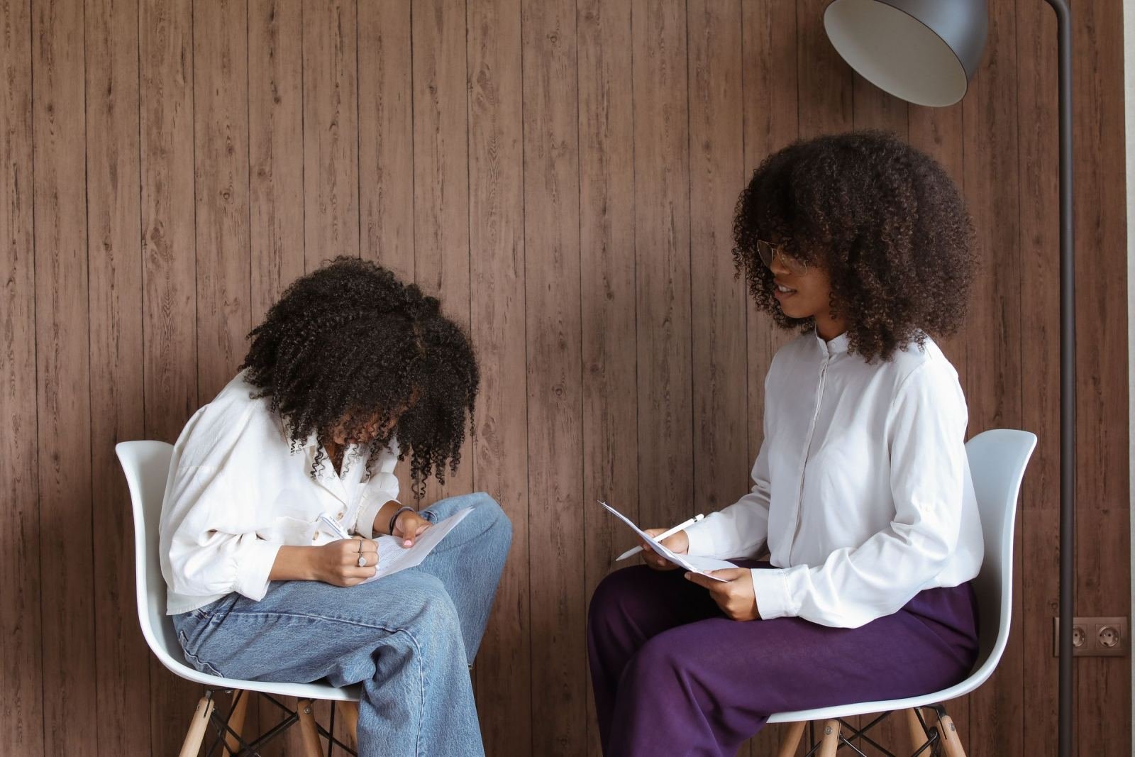 Two women sit in white chairs, one writing on paper and the other holding a clipboard