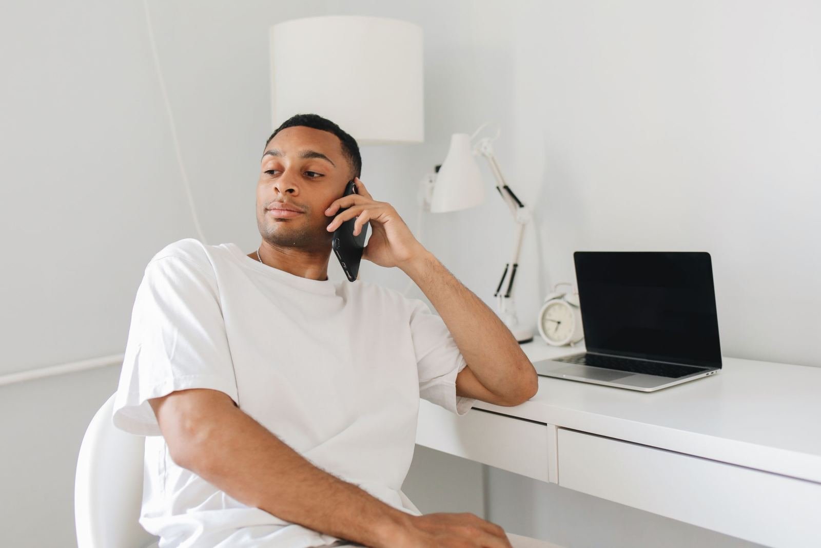 A man in a white t-shirt talks on a mobile phone while sitting at a white desk