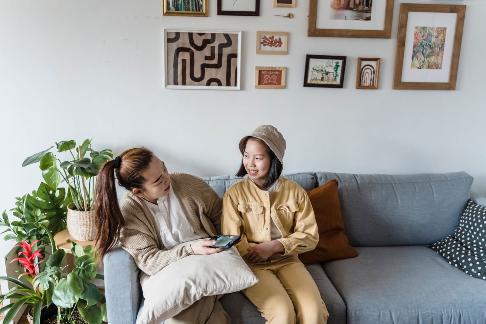 Two women sit on a gray sofa together, one holding a smartphone