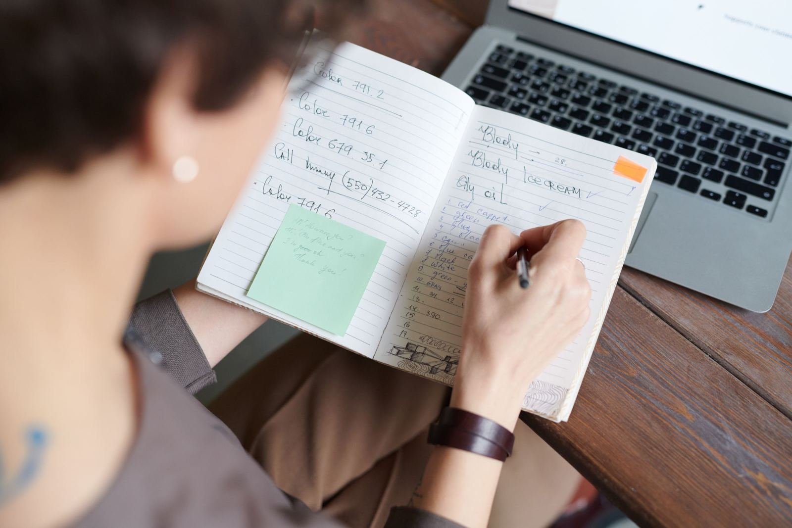Someone writes in a notebook beside a laptop on a wood desk