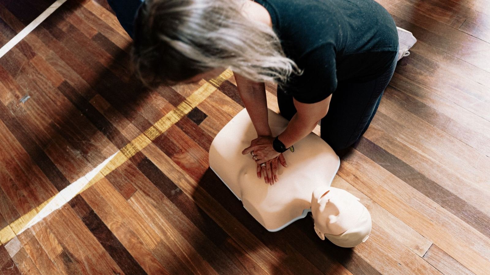 CPR mannequin lies on a wooden floor while someone performs chest compressions.