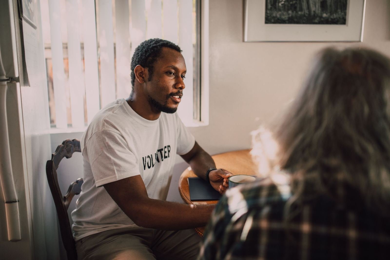 A man in a volunteer t-shirt speaks with another person across a wooden table