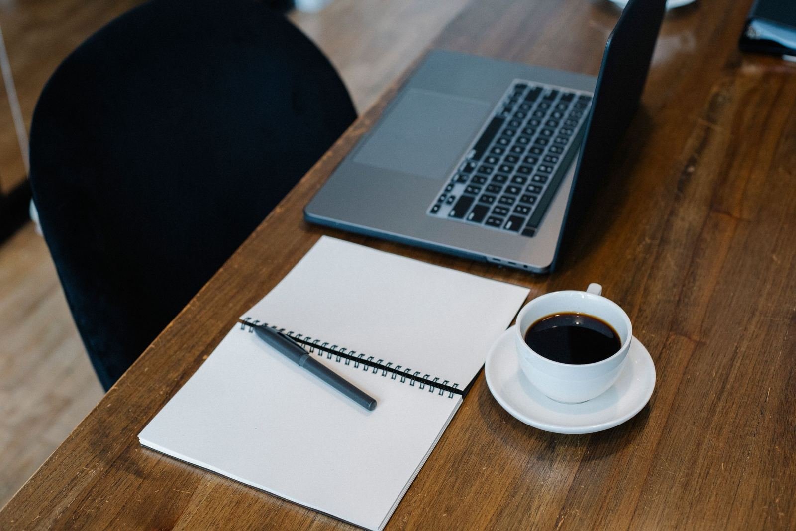 A laptop, notebook, pen, and cup of coffee on a wooden desk